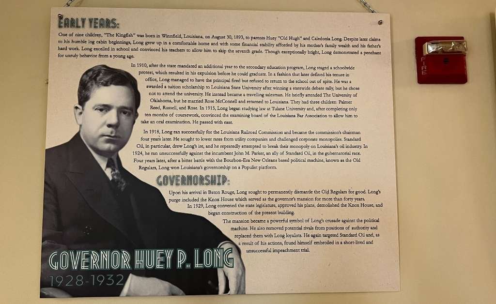 Interpretive museum panel featuring a biography of Governor Huey P. Long (1928–1932), displayed at the Old Governor's Mansion. The panel includes sections titled "Early Years" and "Governorship," with a black-and-white portrait of a young Huey Long in a formal suit. A red fire alarm is visible on the adjacent wall.