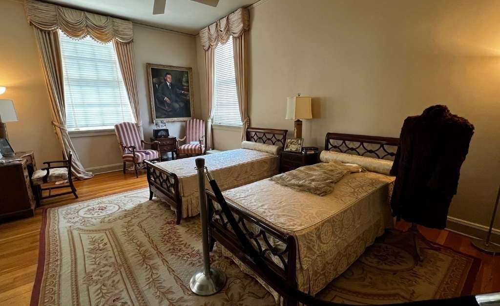 A view of the Earl K. Long Bedroom at the Old Governor’s Mansion, showing twin beds, vintage furniture, a portrait of Earl Long, and a fur coat on display, which hints at his infamous affair.