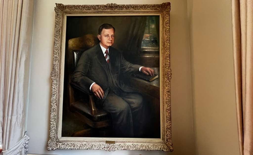 A formal portrait of Governor Huey P. Long seated in a carved wooden chair, wearing a dark suit and holding a book, framed in an ornate gold frame and displayed on a wall at the Old Governor’s Mansion.