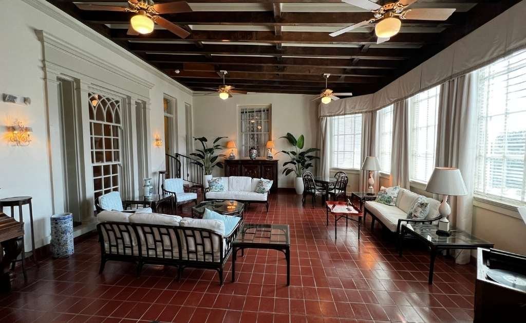 A bright sunroom with terracotta tile flooring, large windows with white curtains, wooden ceiling beams, and a mix of wicker and upholstered seating arranged for conversation.