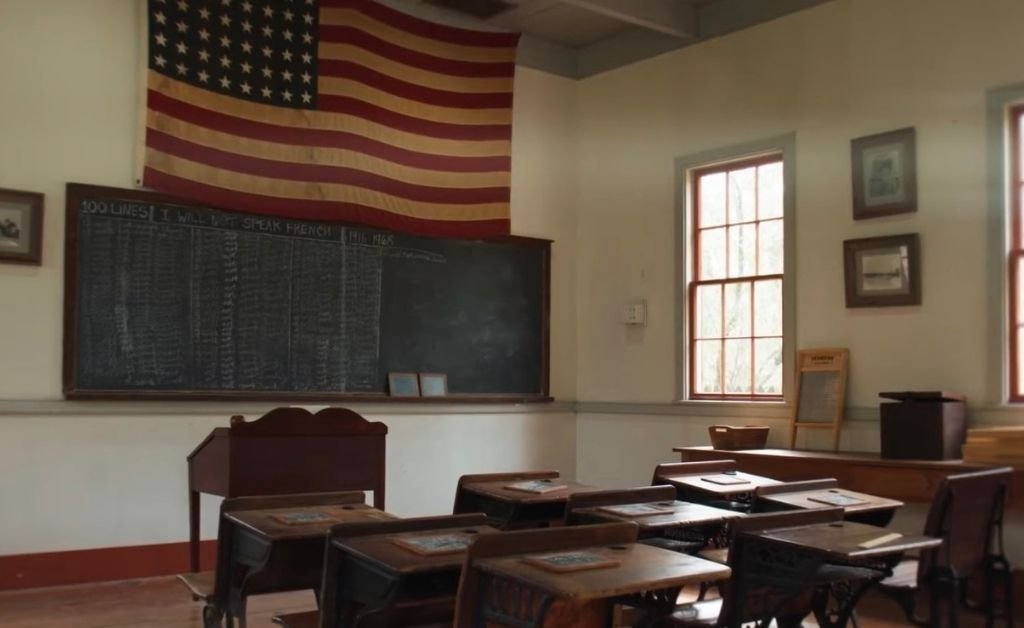 Vintage classroom with wooden desks, chalkboard, and a U.S. flag, showing “I will not speak French” written repeatedly—reflecting language suppression in Louisiana schools.