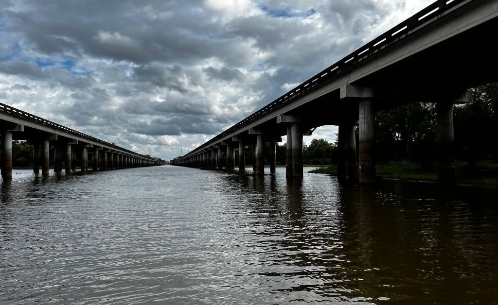 A dramatic view of twin highway bridges spanning the Atchafalaya Basin, taken from the water below, with moody clouds overhead and reflections on the swamp surface.