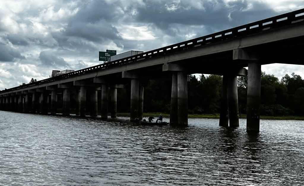 One of the twin spans of the I-10 Atchafalaya Basin Bridge, with a boat passing underneath and the Butte La Rose exit sign visible, under dramatic cloudy skies.