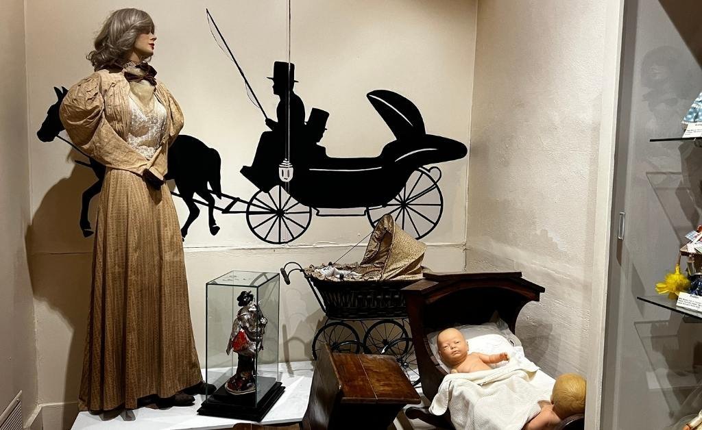Museum display featuring a woman’s period dress, a vintage baby cradle with a doll, and a backdrop illustration of a horse-drawn carriage, representing domestic life in historical Louisiana.