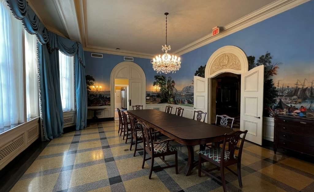 Elegant dining room inside the Old Governor's Mansion featuring a long wooden table, chandelier, arched doorways, blue drapes, and hand-painted French Zuber wallpaper depicting scenic American landscapes.