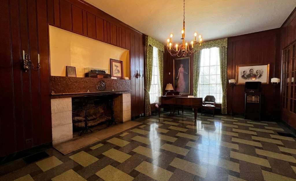 A formal room with dark wood-paneled walls, checkered green-and-black tile floor, a central chandelier, a decorative fireplace, portraits on the walls, and a wooden writing desk by tall windows with green curtains.