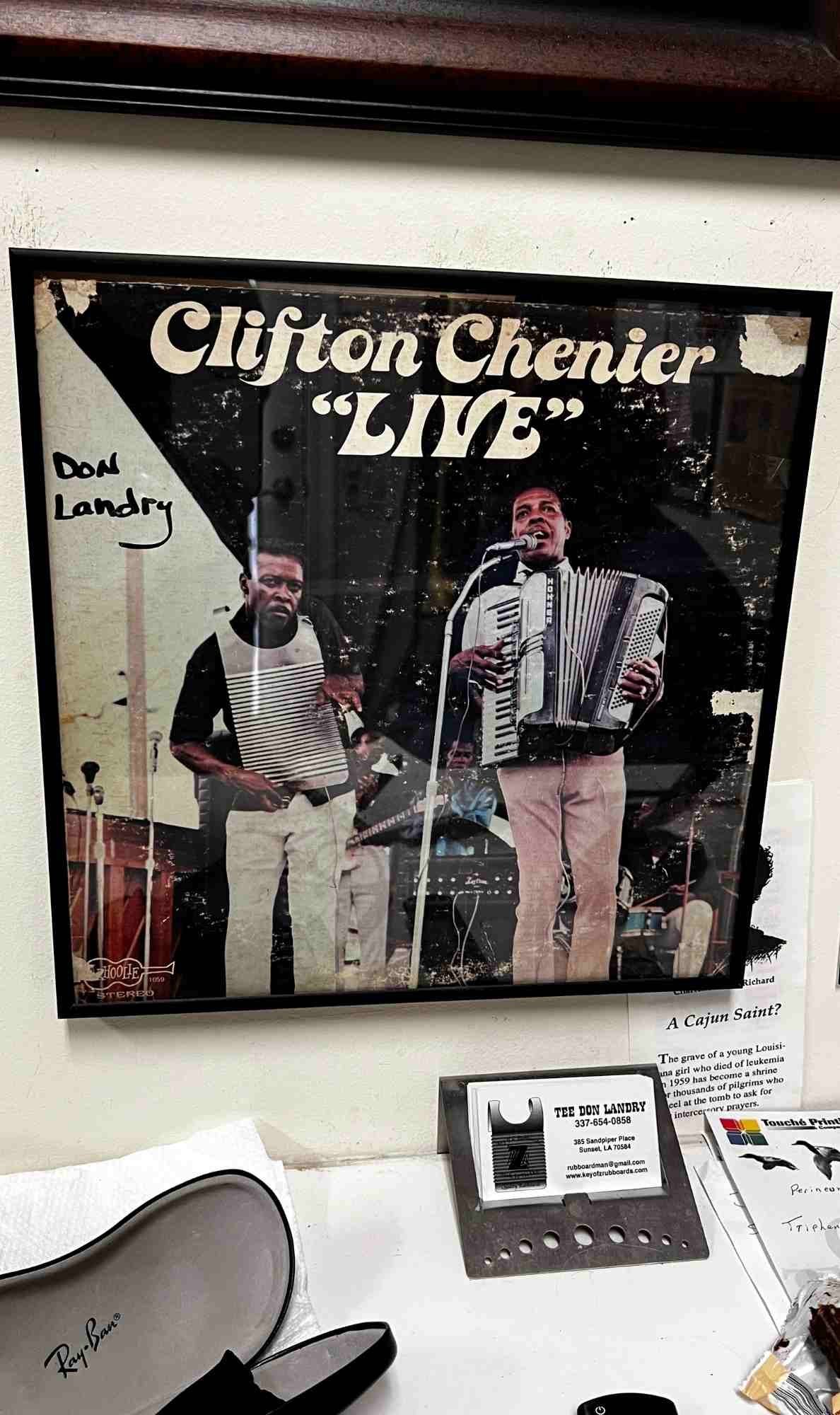 Framed “Clifton Chenier LIVE” album cover displayed above a shelf holding Tee Don Landry’s business card and a small metal rubboard sample.