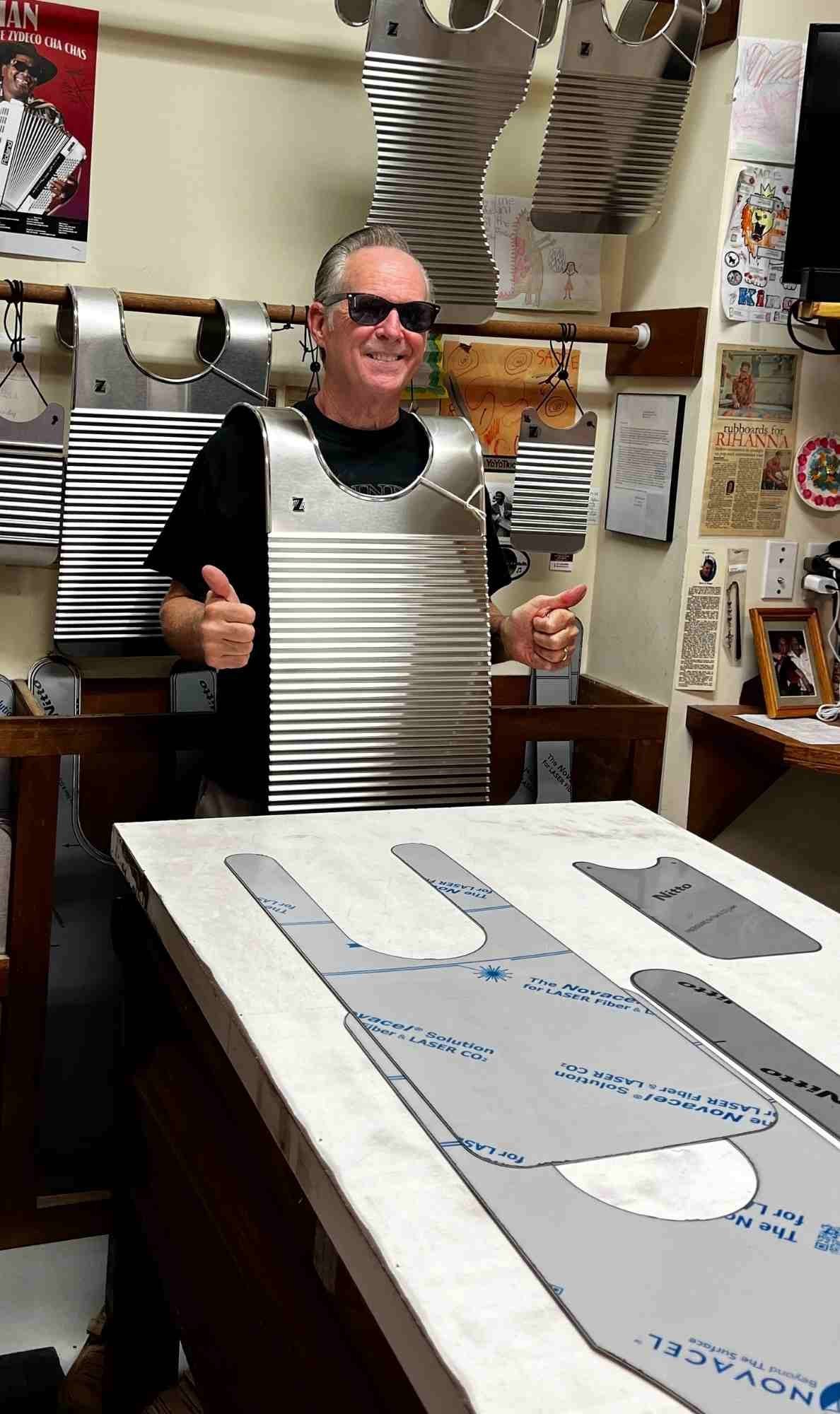 Tee Don Landry smiles and gives two thumbs up while wearing one of his handcrafted stainless steel rubboards inside his workshop, with rubboard templates and tools visible around him.