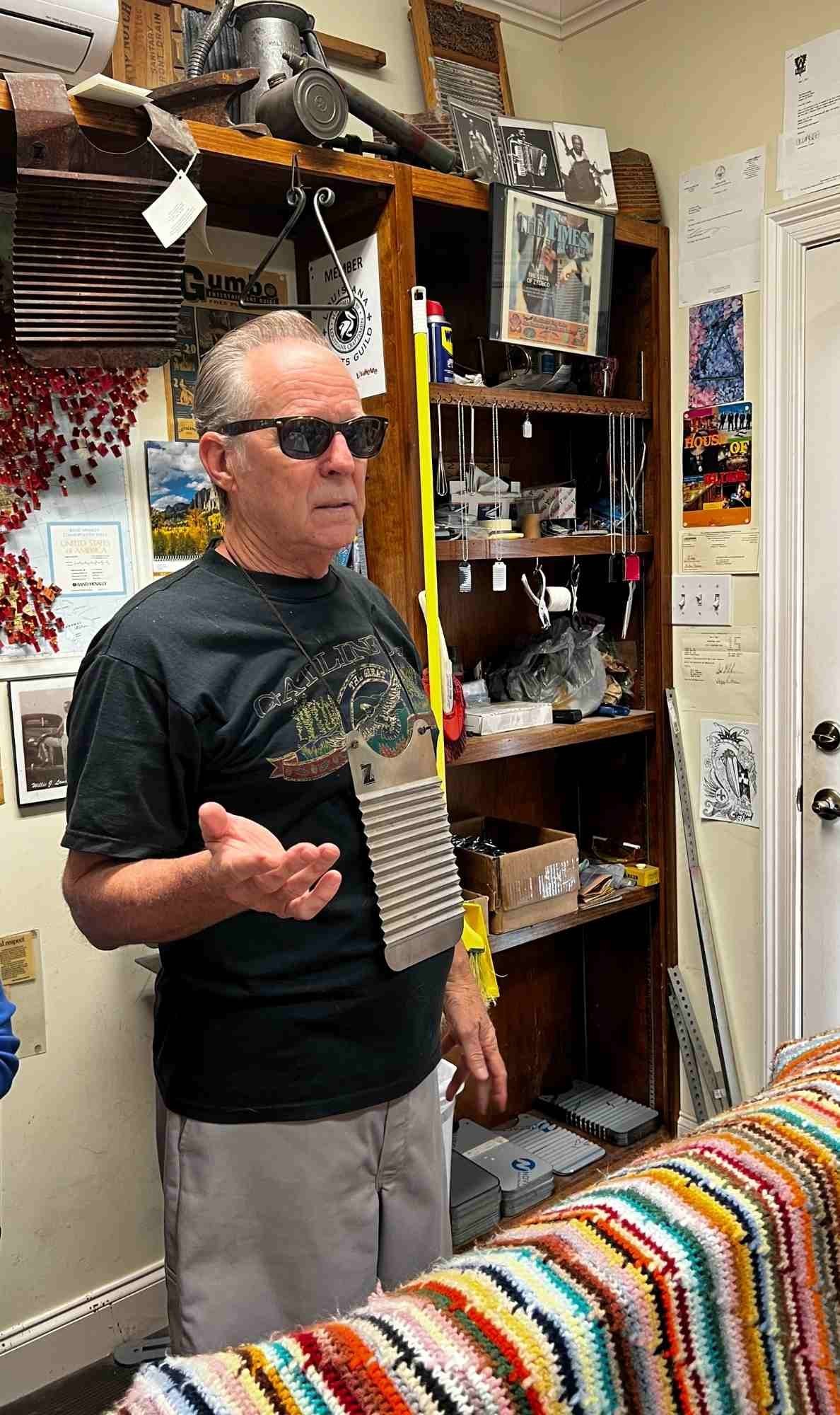 Tee Don Landry explains rubboard craftsmanship in his workshop, wearing a small rubboard pendant around his neck, with tools, memorabilia, and colorful crochet fabric in the background.