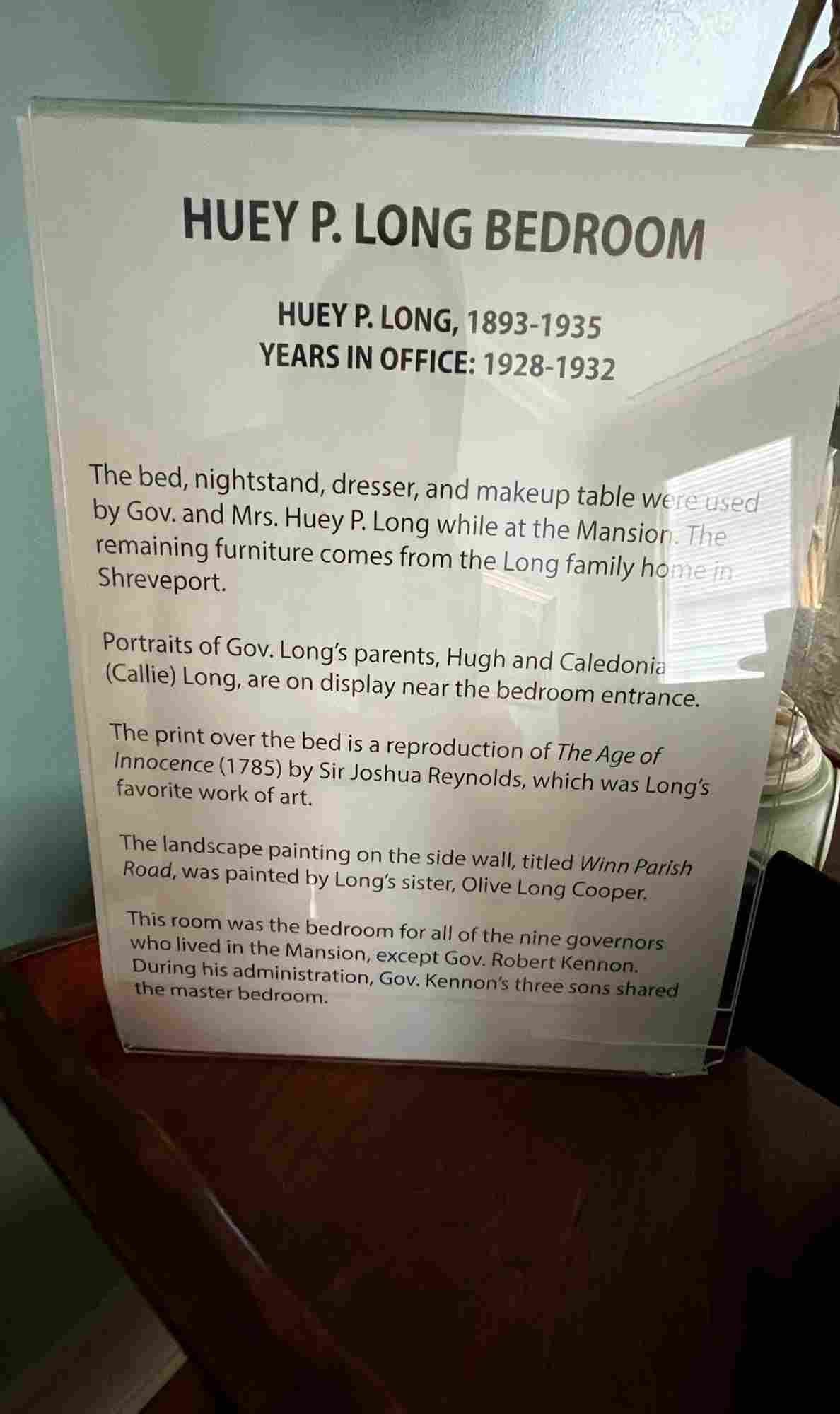 Informational display about the Huey P. Long Bedroom inside the Old Governor's Mansion. The sign provides historical context on furnishings used by Huey and Mrs. Long, portraits of his parents, his favorite artwork, and a landscape painting by his sister. It also notes that this bedroom was used by all nine governors except Robert Kennon.