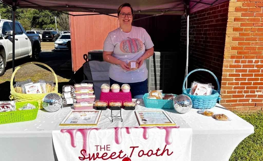 A woman smiles behind a booth for "The Sweet Tooth" at the Sabine Free State Festival, displaying colorful baked goods in baskets and trays under a pink canopy on a sunny day.