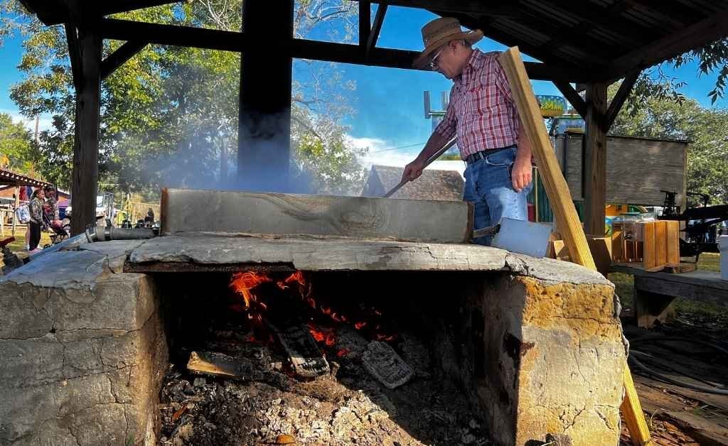 A man in a cowboy hat and plaid shirt stirs a large cast iron kettle over an open fire pit at the Sabine Free State Festival, with smoke rising under a shaded wooden pavilion.
