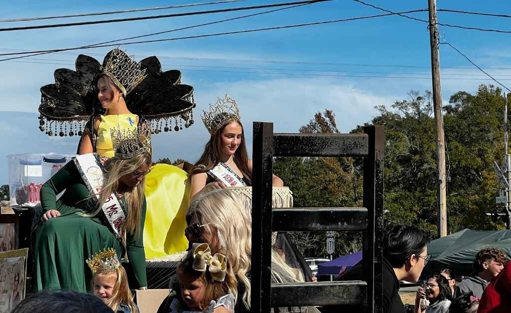 A parade float carrying pageant queens and young girls in crowns and sashes. One queen stands out in a bright yellow dress with an elaborate black fan-shaped headdress, while others wave and smile at the crowd.