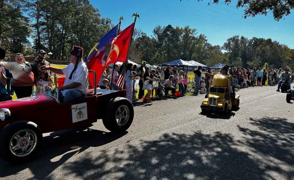 A festive parade scene at the Sabine Free State Festival, featuring a Shriner driving a red miniature car with military emblems and a U.S. Marine Corps flag, followed by a small yellow toy truck, as spectators line the sunny roadside.