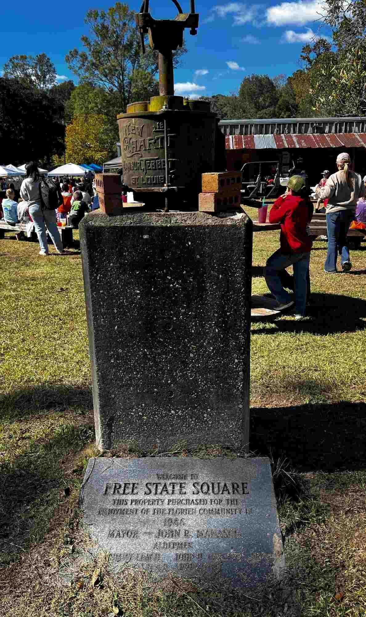 A close-up of the Free State Square monument at the Sabine Free State Festival, featuring a historical iron pump and engraved plaque commemorating the 1986 dedication for community use.