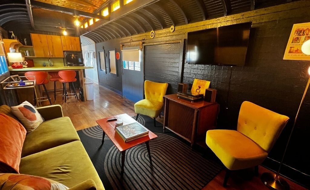 Retro-inspired living room inside the Train Wreck Inn rail car, featuring yellow mid-century chairs, a vintage record console, colorful sofa, and an open kitchen with bold accents.