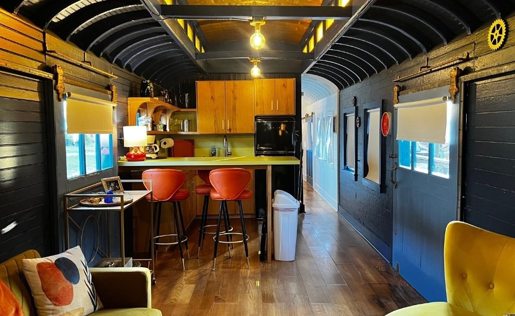 Interior of the Train Wreck Inn rail car featuring a retro kitchen with red bar stools, warm wood cabinetry, and curved black ceilings with yellow accent windows.