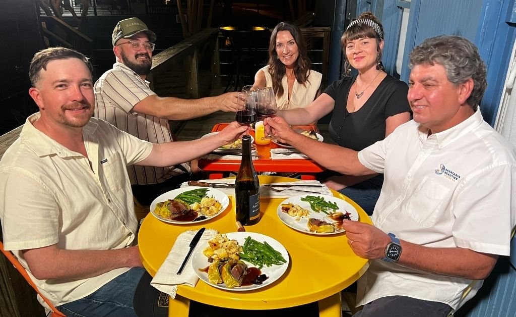 Group enjoying a privately cooked storytelling dinner at the Train Wreck Inn, with Karen and Carly Courville toasting wine at a colorful outdoor table set with plated dishes.