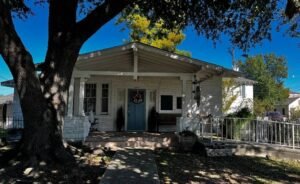 Front exterior of the Clancy House in Kenner, Louisiana, a historic 1922 home with a covered porch, blue front door, and large oak tree in Rivertown.