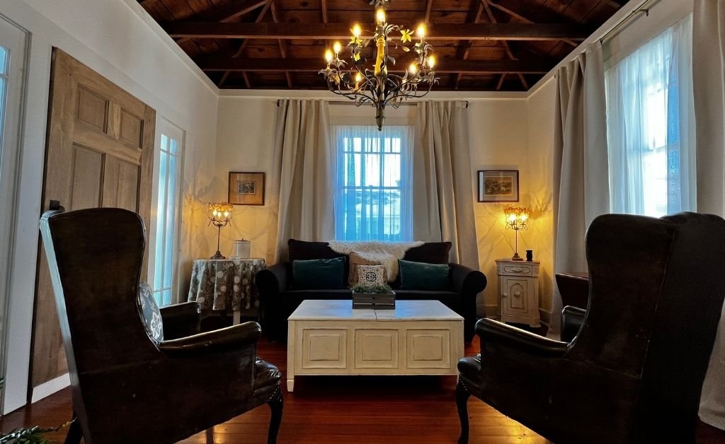 Living room inside the Clancy House featuring vintage furnishings, exposed wood ceiling beams, soft lighting, and a chandelier creating a warm, historic atmosphere.