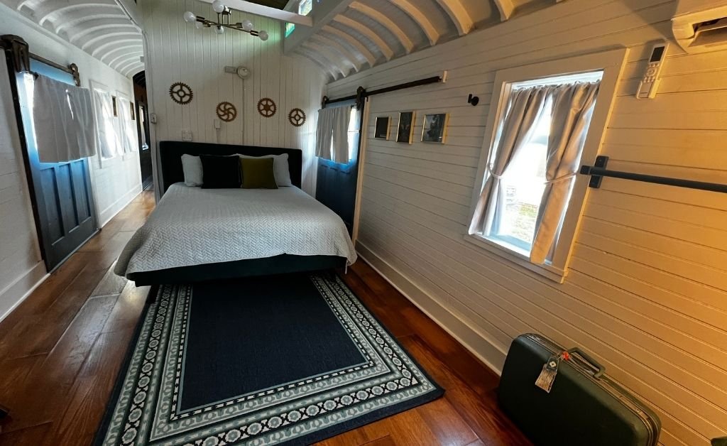 Bedroom inside the Train Wreck Inn rail car featuring a neatly made bed, white paneled walls, gear-shaped decor, soft window curtains, and a vintage suitcase on the floor.