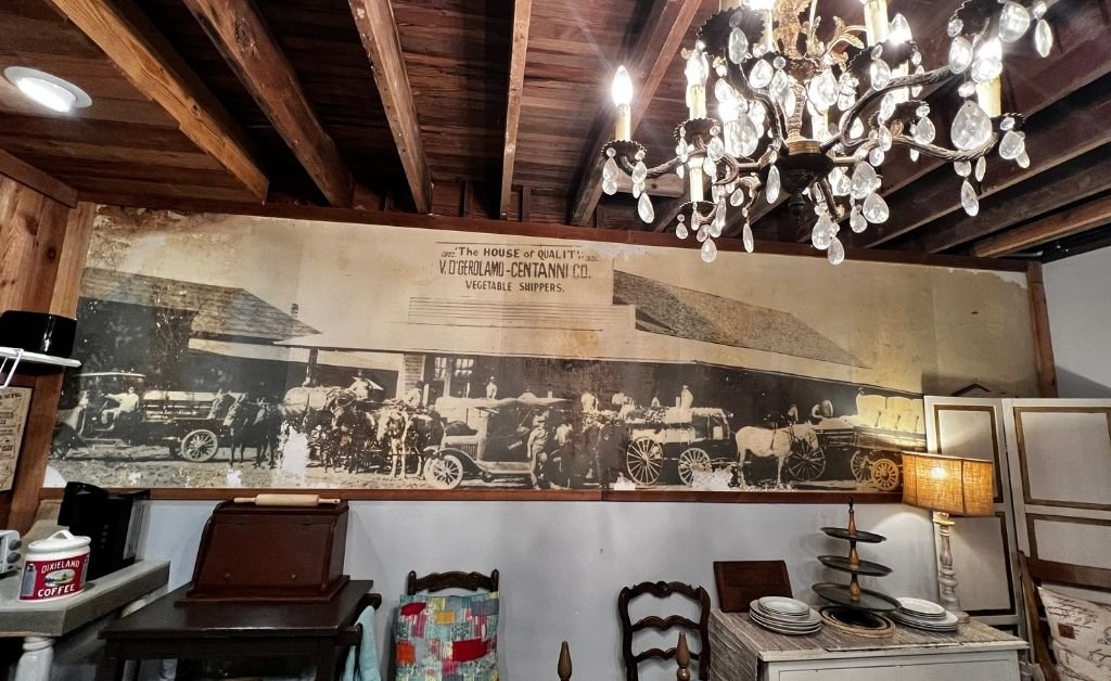 Historic mural inside the Clancy House in Kenner depicting Italian and Irish immigrant vegetable shippers, displayed beneath exposed wooden beams and a crystal chandelier.