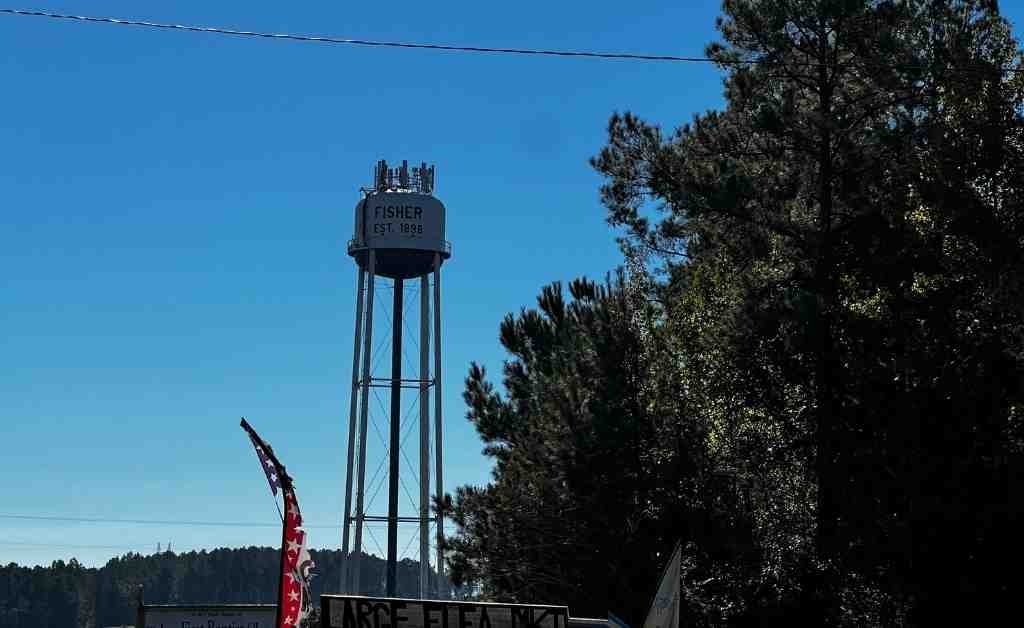 Water tower in Fisher, Louisiana, marked “Est. 1898,” rising above trees against a clear blue sky