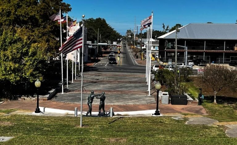 Flag-lined walkway and sculpture overlooking Rivertown Kenner, Louisiana, with Williams Boulevard stretching toward the Mississippi River.