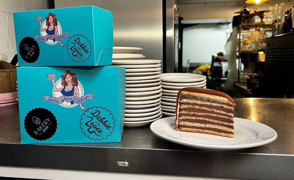 Slice of Doberge cake on a plate beside Debbie Does Doberge bakery boxes at Debbie on the Levee in Kenner.