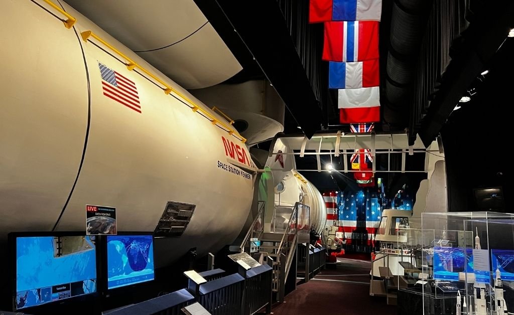 Interior of a NASA exhibit featuring a large space station module labeled “NASA Space Station,” with interactive screens, display cases, and international flags hanging overhead.