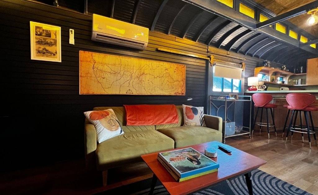 Cozy living area inside the Train Wreck Inn rail car featuring a mid-century sofa, colorful pillows, a vintage map wall art, and a bar counter with red stools.