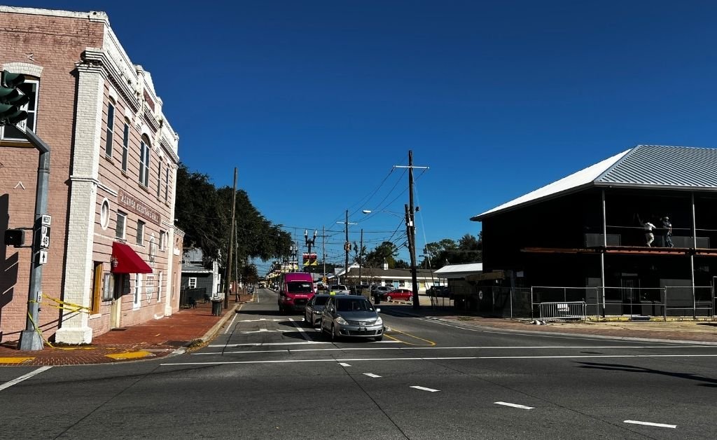 Street view of Kenner, Louisiana, showing the historic Kenner Heritage Hall on the left and a modern riverside structure on the right under a clear blue sky.