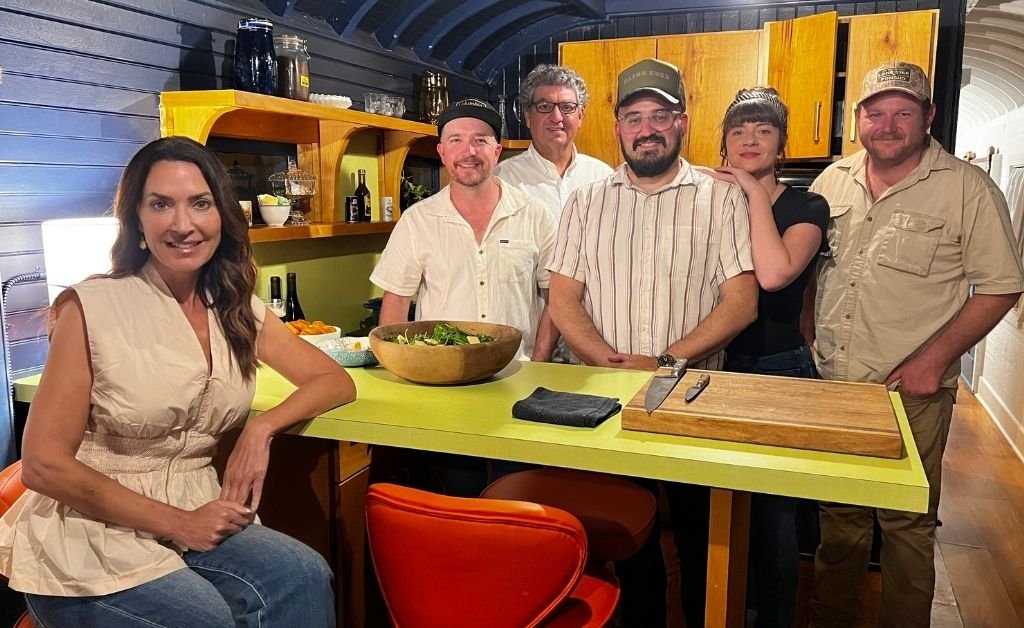 Karen seated at the Train Wreck Inn rail car kitchen counter with Carly Viator Courville and the creative team standing behind her.