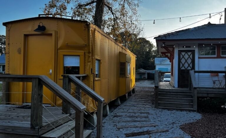 Vintage yellow train car at the Train Wreck Inn in Grand Coteau, Louisiana, with a wooden walkway leading between the rail car and a restored depot building under string lights.