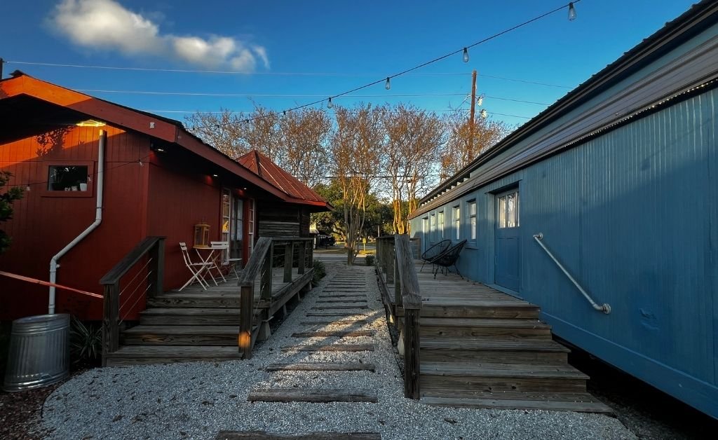 Walkway between the red depot building and the blue vintage rail car at the Train Wreck Inn, with wooden decks, outdoor seating, and string lights overhead in Grand Coteau, Louisiana.