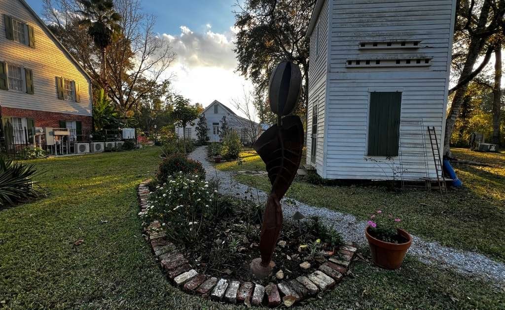 Garden path at Maison Stephanie with historic outbuildings and a metal sculpture set among landscaped beds.