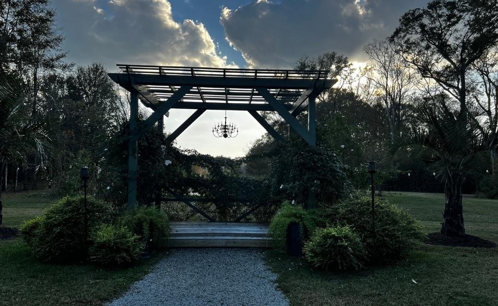 Wooden pergola at Maison Stephanie set among gardens and trees, with a chandelier hanging beneath the structure.