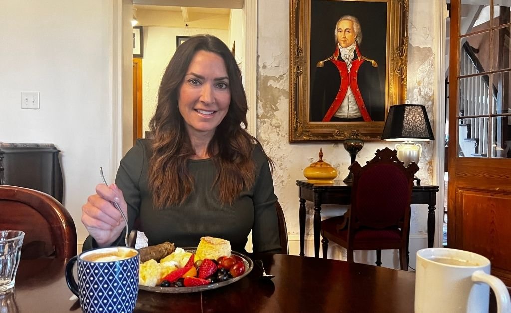Karen LeBlanc enjoying a homemade breakfast inside Maison Stephanie, seated at a historic dining table beneath a framed portrait.