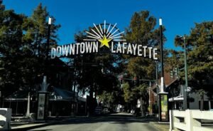 A prominent, stylized metal sign that reads "DOWNTOWN LAFAYETTE" arches over a street lined with trees and buildings in the heart of cajun and creole country.