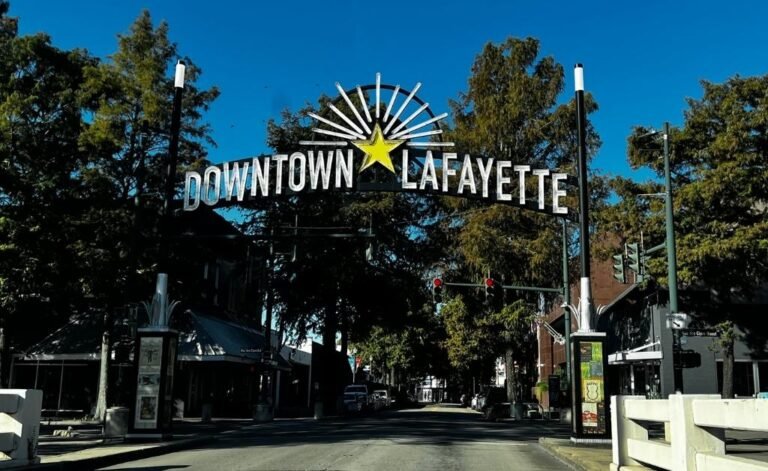 A prominent, stylized metal sign that reads "DOWNTOWN LAFAYETTE" arches over a street lined with trees and buildings in the heart of cajun and creole country.