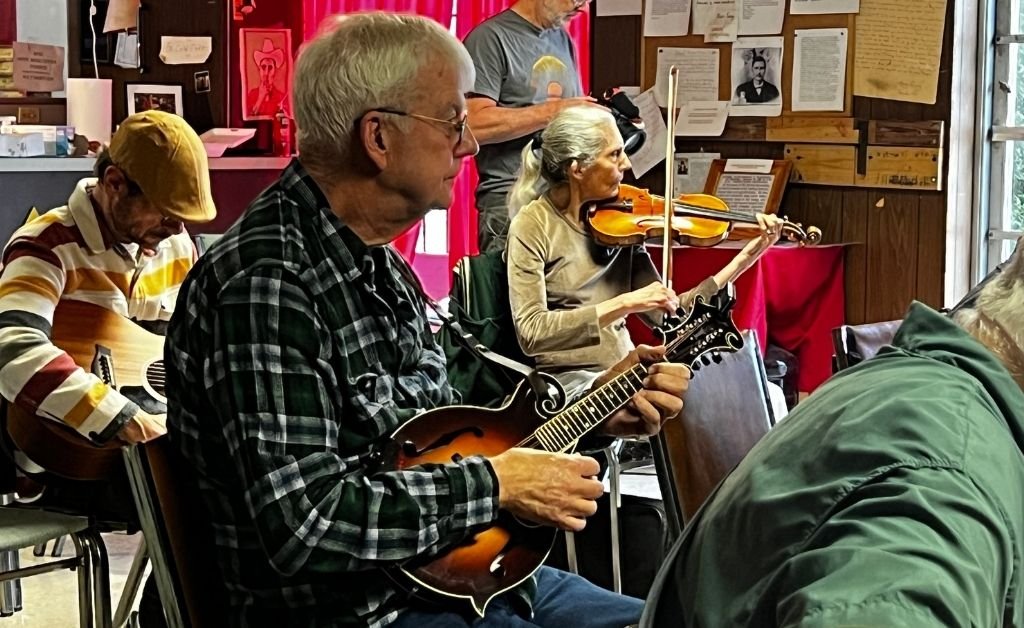 An older man in a green‑plaid shirt plays a mandolin while seated in a chair, focused on his instrument. Behind him, an older woman with gray hair plays a violin, positioned at a music stand in the same indoor setting. Other people sit nearby, and a photographer stands in the background capturing the scene.