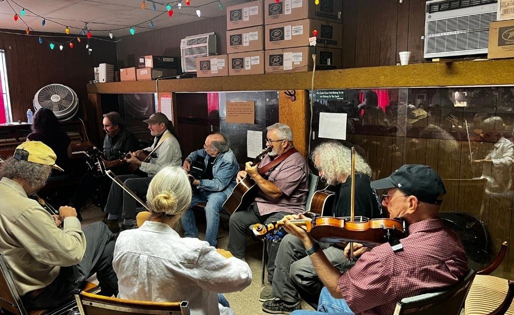 A group of eight musicians are seated in a circle inside the Savoy Music Center, playing various string instruments including violins and guitars in an informal jam session. The wood-paneled room is decorated with colorful string lights and has boxes stacked on a shelf above them. A large glass partition separates the room from an adjacent space.