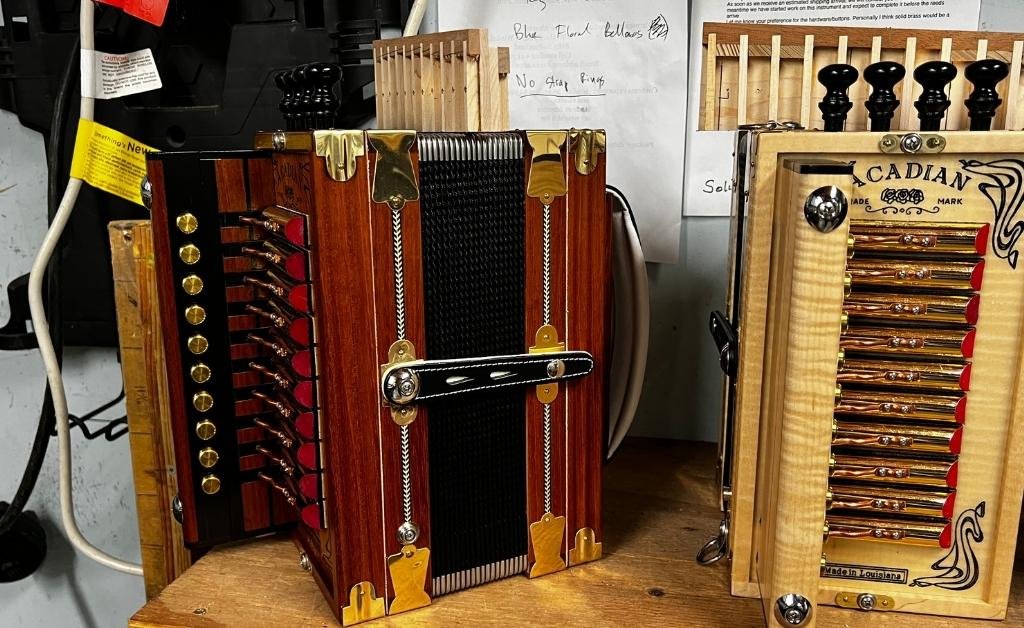 A photograph taken inside the Savoy Family Center shows two accordions placed on a wooden table. The left accordion has a reddish-brown wooden case with brass fittings and black bellows, while the right accordion has a light-colored wood case with gold reeds and the inscription “ACADIAN MARK” on its grille. A handwritten note on the wall behind them mentions “Blue Fluted Bellows” and “No Stop Rings.”