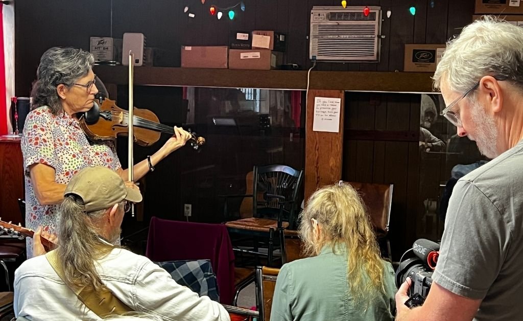 A woman with gray hair plays a violin in an indoor setting at the Savoy Family Center. She stands facing three audience members seated in chairs, one of whom holds a guitar, while another person on the right holds a video camera recording the performance. The room has wooden walls, an air conditioner, and colorful string lights above a shelf.