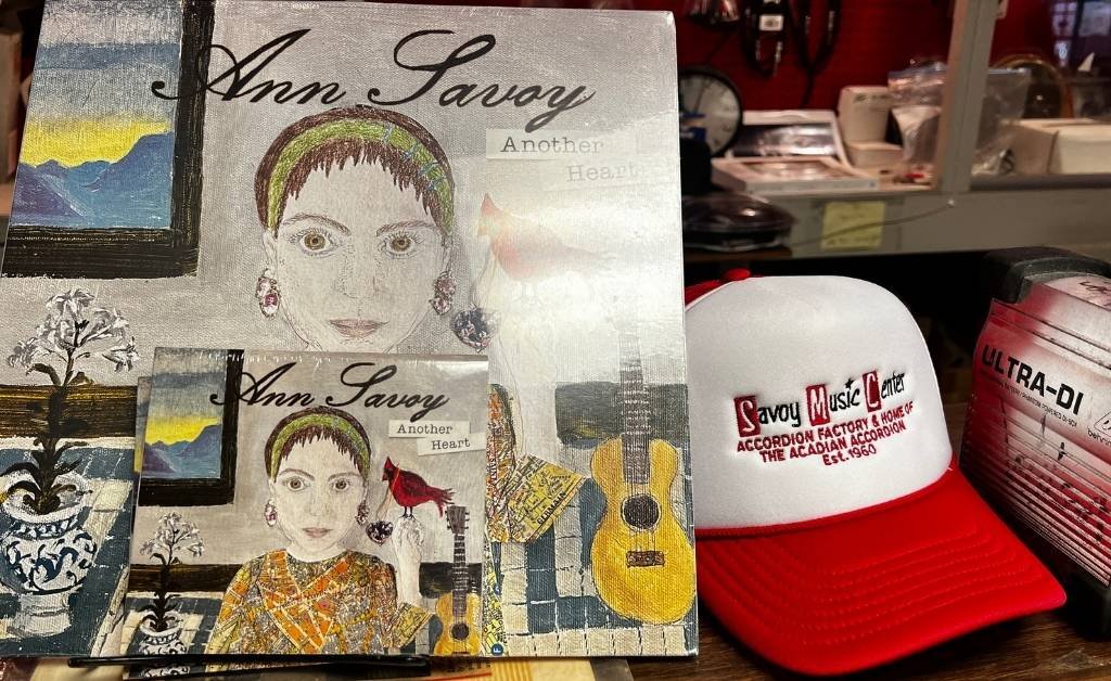 A photograph taken inside the Savoy Family Center shows two mixed‑media artworks featuring a portrait of Ann Savoy with the phrase “Another Heart,” accompanied by painted guitars and other decorative elements. Beside the artworks rests a red‑brimmed white trucker hat with red embroidered text reading “Savoy Music Center ACCORDION FACTORY & HOME OF THE ACADIAN ACCORDION Est. 1960.” A pink “ULTRA-DI” canister is partially visible on the right.