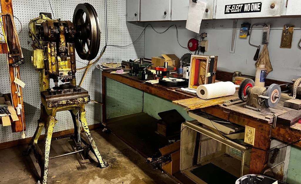 A workshop scene showing a yellow, worn industrial machine with a large flywheel on a stand next to a long wooden workbench cluttered with tools, a paper towel roll, grinding wheels, and various workshop supplies. The wall behind has pegboard, cabinets, and a “RE‑ELECT NO ONE” sticker.