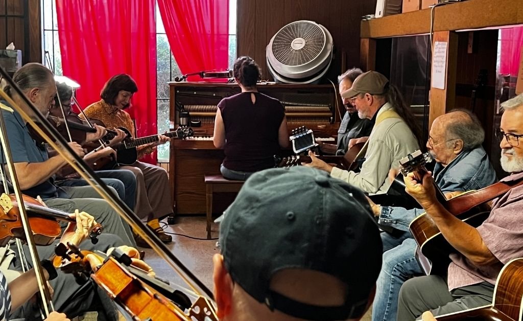 A group of musicians rehearses in the Savoy Family Center. Eight men sit in a circle playing guitars and a violin while a woman with her back to the camera plays an upright piano positioned in front of a large window with red curtains and a fan on top. The setting shows a cozy, wood‑paneled music room.