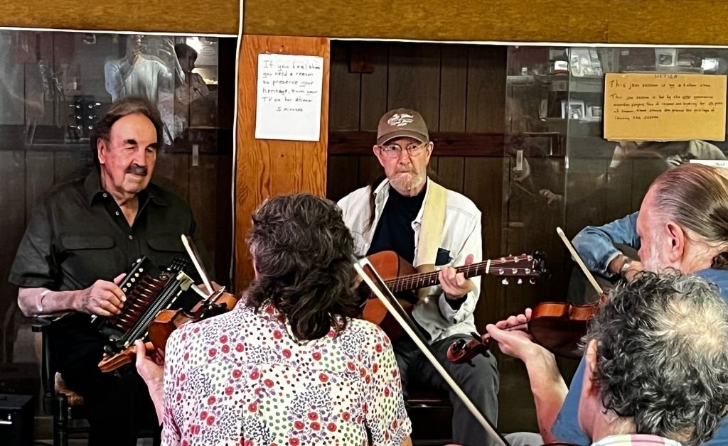 The image shows an interior scene at the Savoy Family Center where two musicians are performing for two audience members holding violins. On the left, Marc Savoy, wearing a black shirt, plays an accordion, while a man in a white jacket and brown cap plays an acoustic guitar beside him. The setting includes wooden walls with handwritten notices visible in the background.