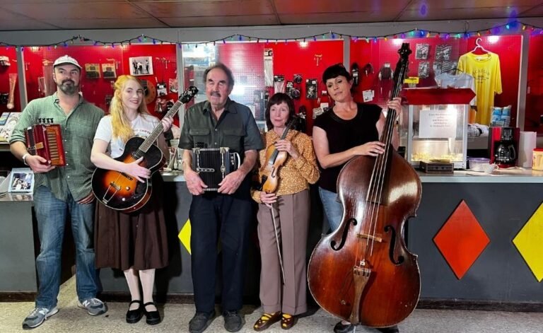 The Savoy family, five musicians, stand in a row inside a music shop with red walls adorned with colorful string lights and merchandise. From left to right, they hold a washboard, an electric guitar, an accordion, a violin, and an upright double bass. The setting includes a counter with diamond-shaped orange and yellow panels and displayed items.
