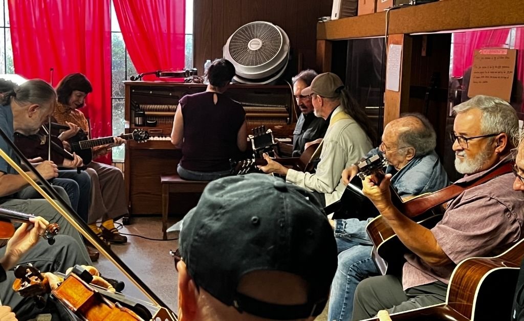 A group of musicians rehearses in the Savoy Family Center. Eight men sit in a circle playing guitars and a violin while a woman with her back to the camera plays an upright piano positioned in front of a large window with red curtains and a fan on top. The setting shows a cozy, wood‑paneled music room.